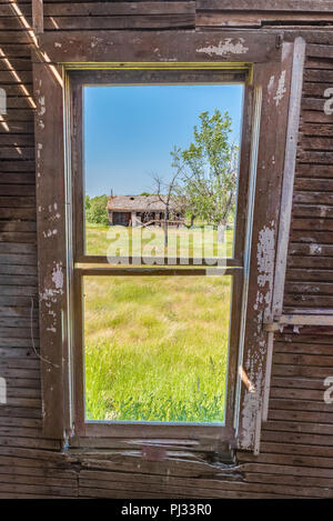 View through the window of an abandoned prairie farmhouse looking out on an old barn surrounded by tall grass Stock Photo