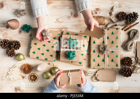 Two small children's hands holding out gift tag, bow and present for decorating wrapped into brown paper Christmas presents, pine cones, baubles, hess Stock Photo