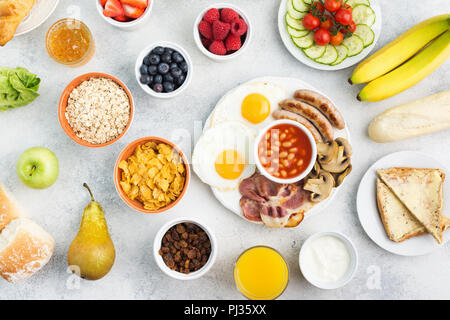 English breakfast fried eggs, sausages, bacon and mushrooms with selection of fruits and vegetables, breads and juice on the grey white table, top vie Stock Photo