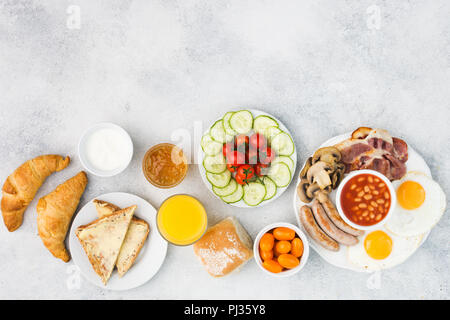 Delicious breakfast fried eggs, sausages, bacon and mushrooms with selection of vegetables, croissants and juice on the grey white table, top view, co Stock Photo