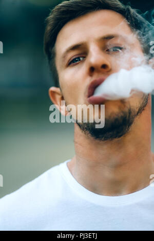 Modern young man with a beard fun Vaporizers Outdoor. Male in white t-shirt vape ecig. dark background, the evening sunset over the city. toned image. Stock Photo