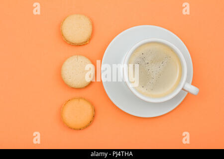 White porcelain cup of coffee on saucer and delicious macarons cakes on peach colored background. Top view Stock Photo