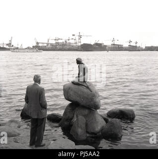 1950s, historical, Copenhagen, Denmark, at the waterside, by the shore of the Langeline, a gentleman looking at 'the Little Mermaid', a bronze statue on a rock by Edvard Eriksen, depicting a mermaid becoming human. Although small in size, it is one of the city's most famous tourist attractions, and based on a character from a fairy tale by Danish author Hans Christian Andersen. Stock Photo