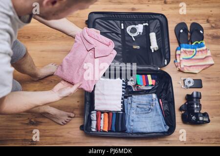 Preparing for trip. Young man packing clothing for vacation, flip flop, camera and other things on hardwood floor. Stock Photo