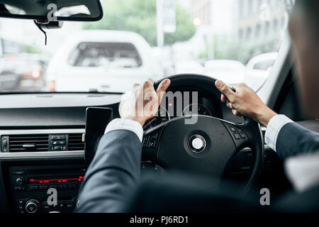 Rear view of man in formal clothes holding the steering wheel of his car with both hands. Businessman driving car with mobile phone clipped to dashboa Stock Photo