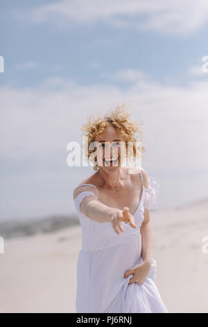Happy woman tourist enjoying on the beach. Smiling woman in white gown running on beach reaching out for something. Stock Photo