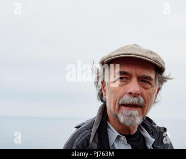 Close up profile of thoughtful senior man with a gray beard wearing a cap gazing into distance, faint ocean horizon in background Stock Photo