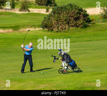 Senior man with golf cart swinging golf club to hit golf ball on golf course in sunshine, Edinburgh, Scotland, UK Stock Photo