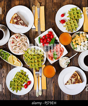 Breakfast food table. Festive brunch set, meal variety with spinach waffles, salmon, cheese, olives, chicken rolls and cheesecake. Top view. Flat lay Stock Photo