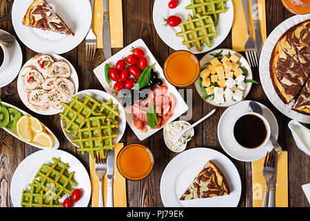 Breakfast food table. Festive brunch set, meal variety with spinach waffles, salmon, cheese, olives, chicken rolls and cheesecake. Top view. Flat lay Stock Photo