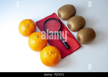 Three mandarins against three kiwis with black magnifying glass on red napkin isolated over white background. Stock Photo
