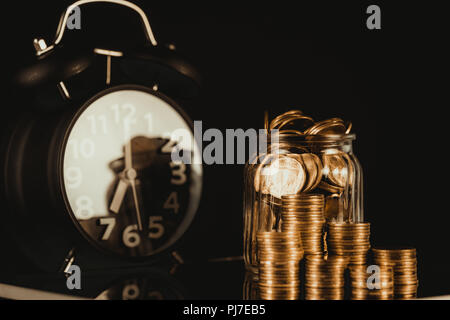 Coins stacks with coin in glass jar bottle and alarm clock in dark room, business and finance concept idea. Stock Photo