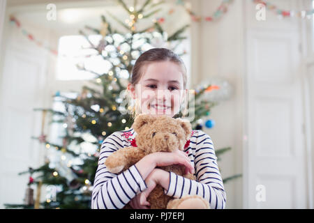 Portrait smiling, cute girl holding teddy bear in front of Christmas tree Stock Photo