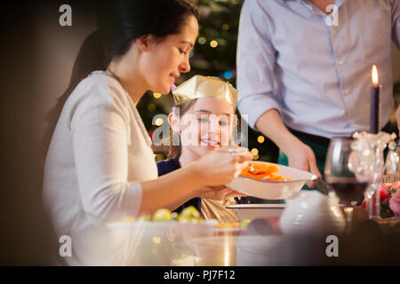 Mother serving carrots to daughter in paper crown at Christmas dinner Stock Photo