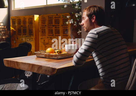 Man sitting and drinking enjoying tea or coffee in a kitchen at home. Pensive, thoughtful young male , view from the shoulder, rear view. Stock Photo