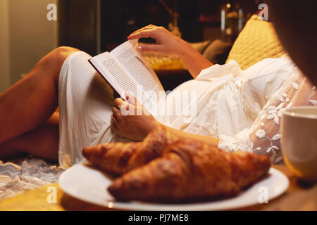 Pregnant woman lying on the bed at bedroom and reading a book about maternity or chicklit, womans novel at home, focus on a belly and book. Lifestyle, Stock Photo