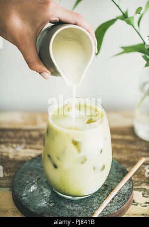 Iced matcha latte drink in glass with coconut milk pouring from pitcher by woman's hand, white wall and plant branches at background, close-up. Summer Stock Photo