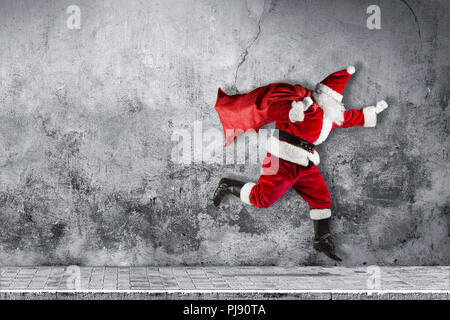 late Santa claus in a hurry with traditional red white costume and bag full of presents running jumping abstract funny christmas xmas concrete backgro Stock Photo
