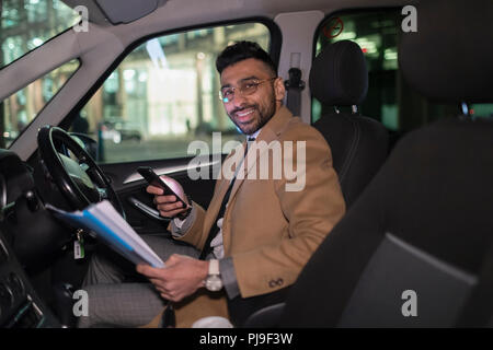 Portrait smiling businessman with smart phone reading paperwork in car at night Stock Photo