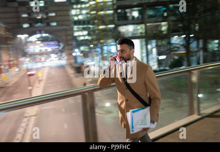 Businessman talking on smart phone, walking on urban pedestrian bridge at night Stock Photo