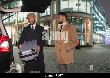 Businessmen loading suitcase into car on urban street corner at night Stock Photo