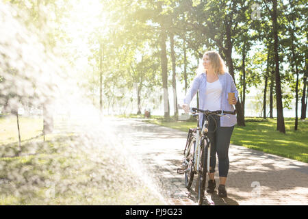 Young blonge woman walking her bicycle in the park and holding a cup of coffee in her hand. Stock Photo