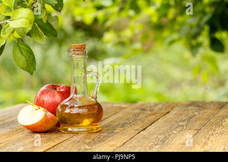Apple vinegar in glass bottle with cork and fresh red apples on old wooden boards with blurred green natural background Stock Photo