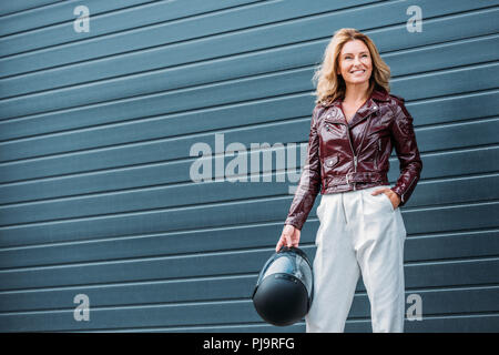attractive woman in leather jacket holding motorcycle helmet on street Stock Photo