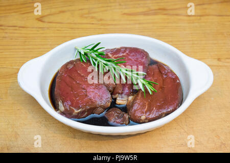 Meat marinating: Beef steak eye fillet in white dish of soy sauce marinade and rosemary herb sprig on wooden background with copy space. Lean cut with Stock Photo