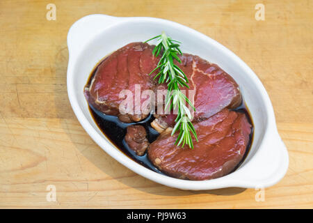 Meat marinating: Beef steak eye fillet in white dish of soy sauce marinade and rosemary herb sprig on wooden background - diagonal composition. Lean c Stock Photo