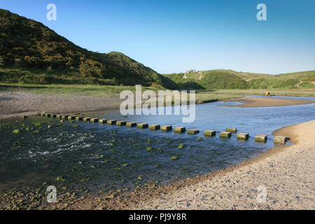Stepping stones cross the stream at Three Cliffs Bay on the Gower peninsula near Swansea, Wles, UK, with Pennard Castle on the hillside in the backgro Stock Photo