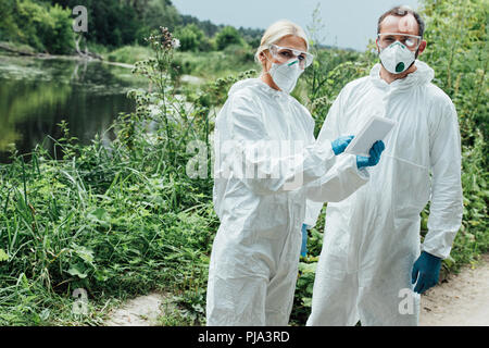 female and male scientists in protective masks and suits working with digital tablet and looking at camera outdoors Stock Photo