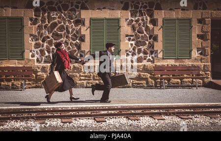 Young couple with vintage suitcase running fast outside a train station to catch the last train for journey. Stock Photo