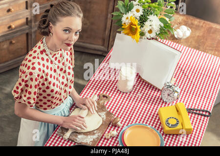 high angle view of beautiful adult housewife kneading dough with rolling pin and looking at camera at kitchen Stock Photo