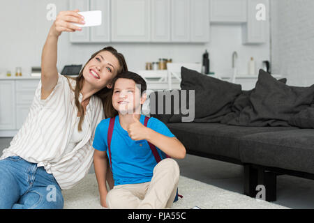 smiling mother and son with backpack and thumb up taking selfie on smartphone together at home on first school day Stock Photo