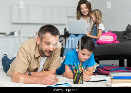 selective focus of father and son doing homework together while mother and daughter using laptop on sofa at home Stock Photo