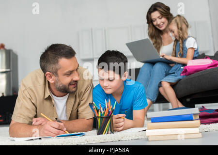 selective focus of man helping son with homework while mother and daughter using laptop on sofa together at home Stock Photo