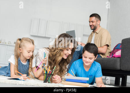 smiling mother helping kids with homework while man using laptop on sofa at home Stock Photo