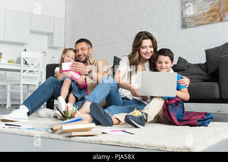 smiling father and daughter taking selfie on smartphone while mother and son using laptop together at home Stock Photo