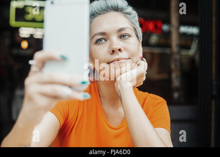 Close up of attractive blonde girl in orange T-shirt making selfie at cafe and drinking coffee. Stock Photo