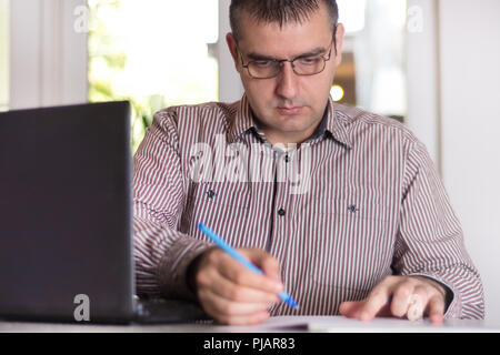 Business man working at modern office with documents and laptop on his desk. Man wears shirt and glass. Blurred windows in background. Close up Stock Photo