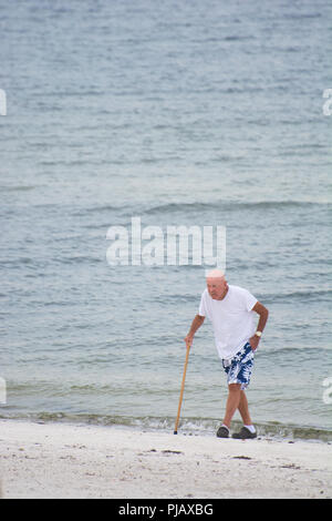 An elderly bald man in brightly colored shorts walks with a cane on the beach at Anna Maria Island, Florida, USA Stock Photo