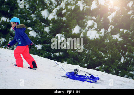 Little girl with security helmet and googles going up on a hill pulling a plastic  Children play outdoors in snow. Kids sled in Alps mountains in wint Stock Photo