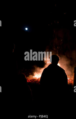 silhouettes of people around a large bonfire underneath a nearly full moon on a winter’s night in the Cevennnes, France Stock Photo