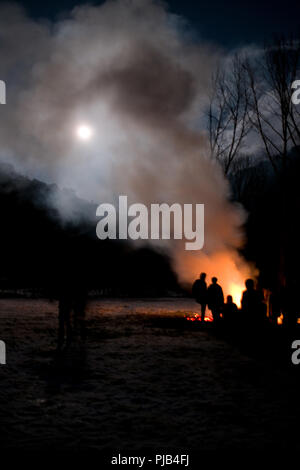 artistic shot of group of people silhouhetted against a bonfire whose smoke drifts up across the sky obscuring full moon on a dark Winter’s night Stock Photo