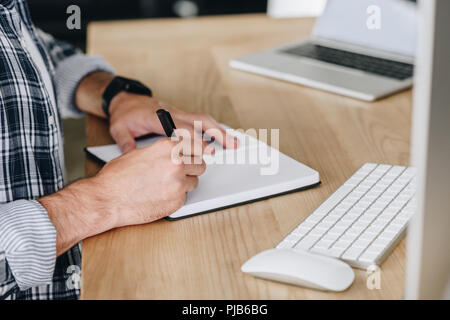 cropped shot of man taking notes in notepad while using desktop computer at workplace Stock Photo