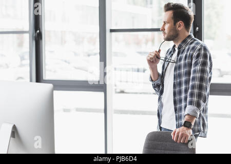 pensive young man holding eyeglasses and looking away while standing at workplace Stock Photo