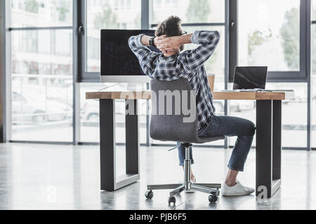 back view of young programmer sitting with hands behind head at workplace Stock Photo