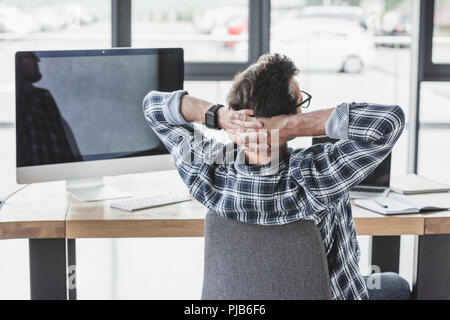 back view of programmer sitting with hands behind head at workplace Stock Photo