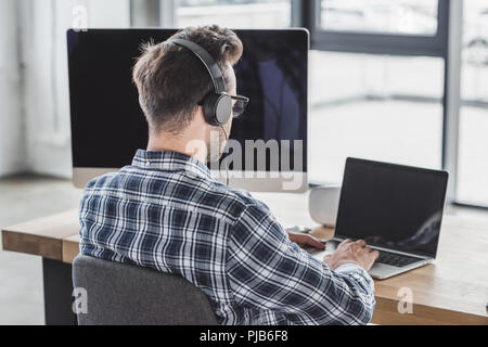 back view of young programmer in headphones and eyeglasses working with laptop Stock Photo